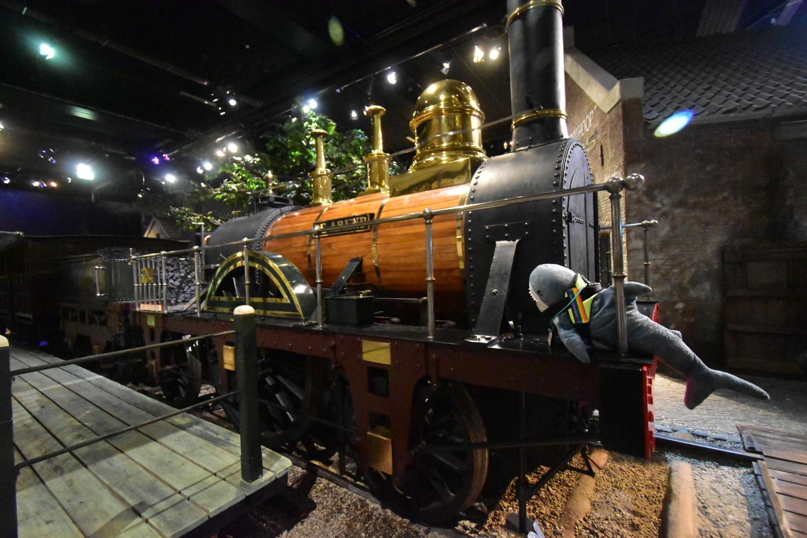A Blåhaj cuddly shark sitting on the front of a replica of 'de Arend', the first steam engine to ride in the Netherlands. The Blåhaj wears a haj-vis harness with rainbow patches and a lanyard with a Dutch OV-chipkaart on it.

You are right, the world would be a more cuddly place with hajar in power!