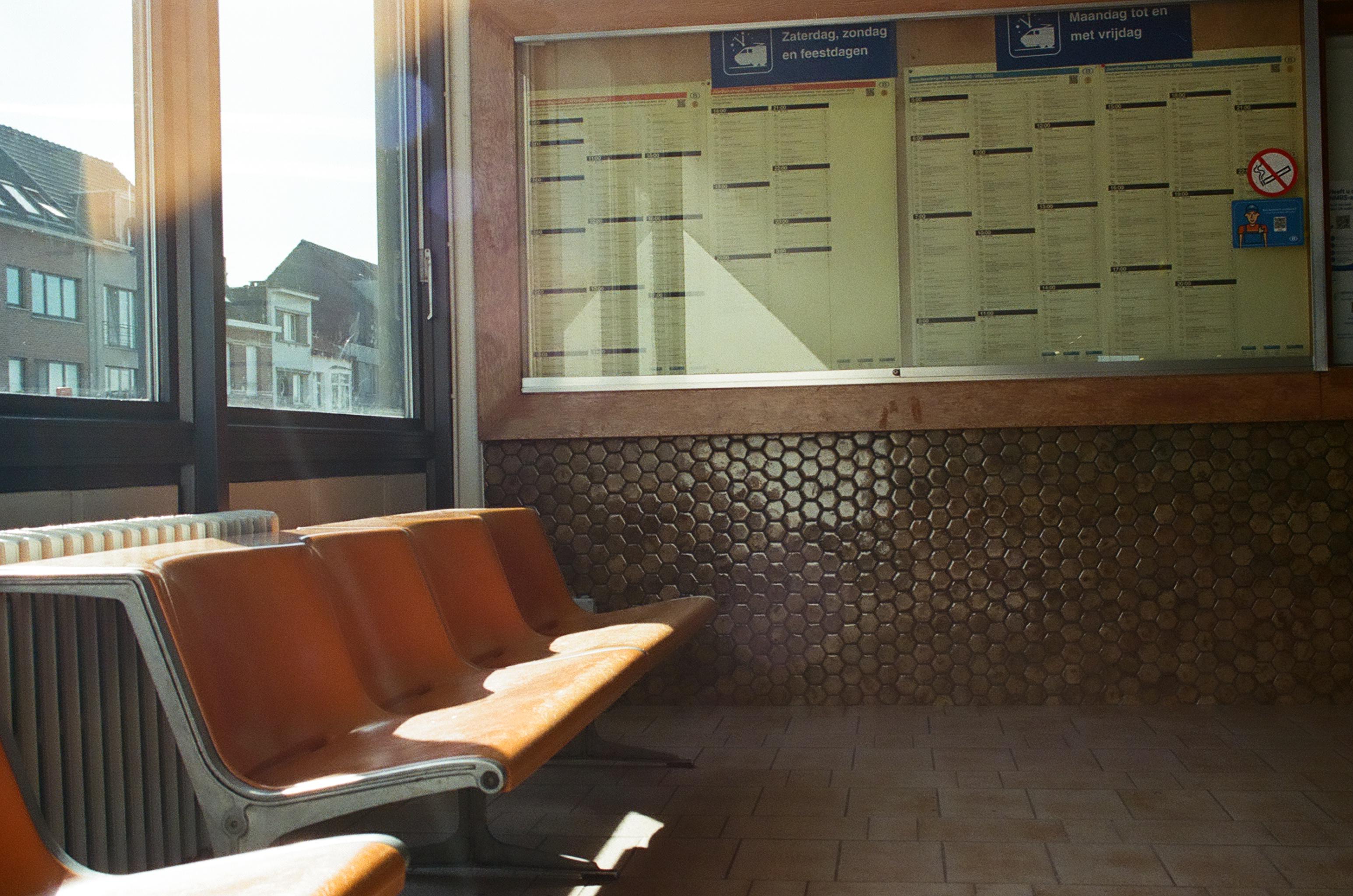 This image shows a row of orange chairs in a waiting area with a window behind them. The window displays a board with text and a sign.